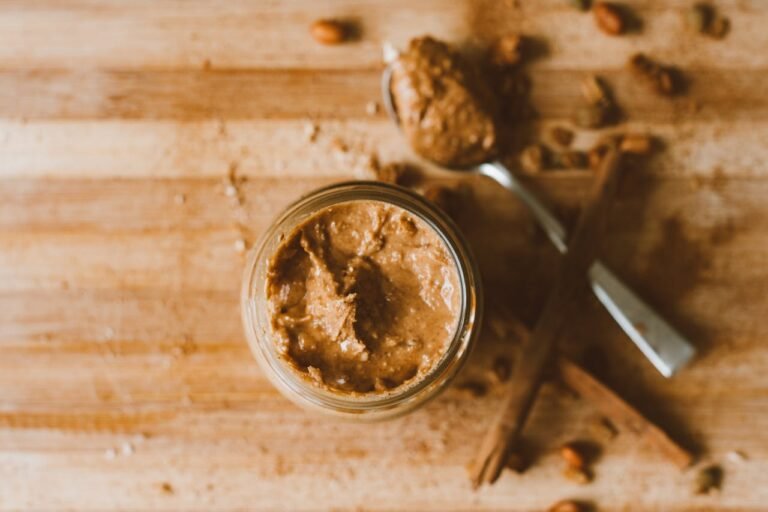 Pot de beurre de cacahuètes crémeux sur une planche en bois avec une cuillère et des bâtons de cannelle en arrière-plan.